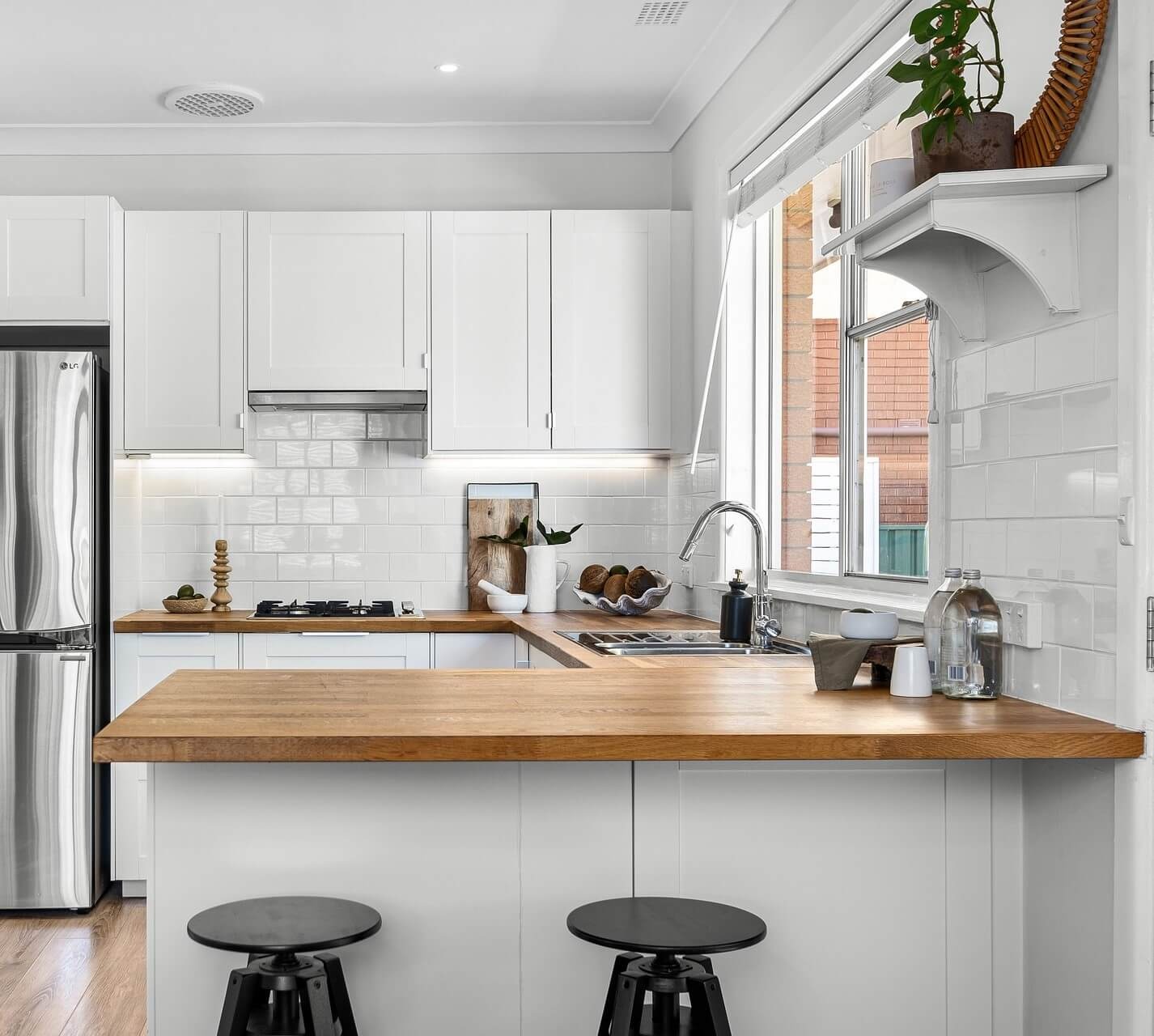 Plan de travail de cuisine avec évier rond en inox, robinet mural chromé, crédence en carreaux blancs et meubles en bois clair, éclairé par une fenêtre avec stores.