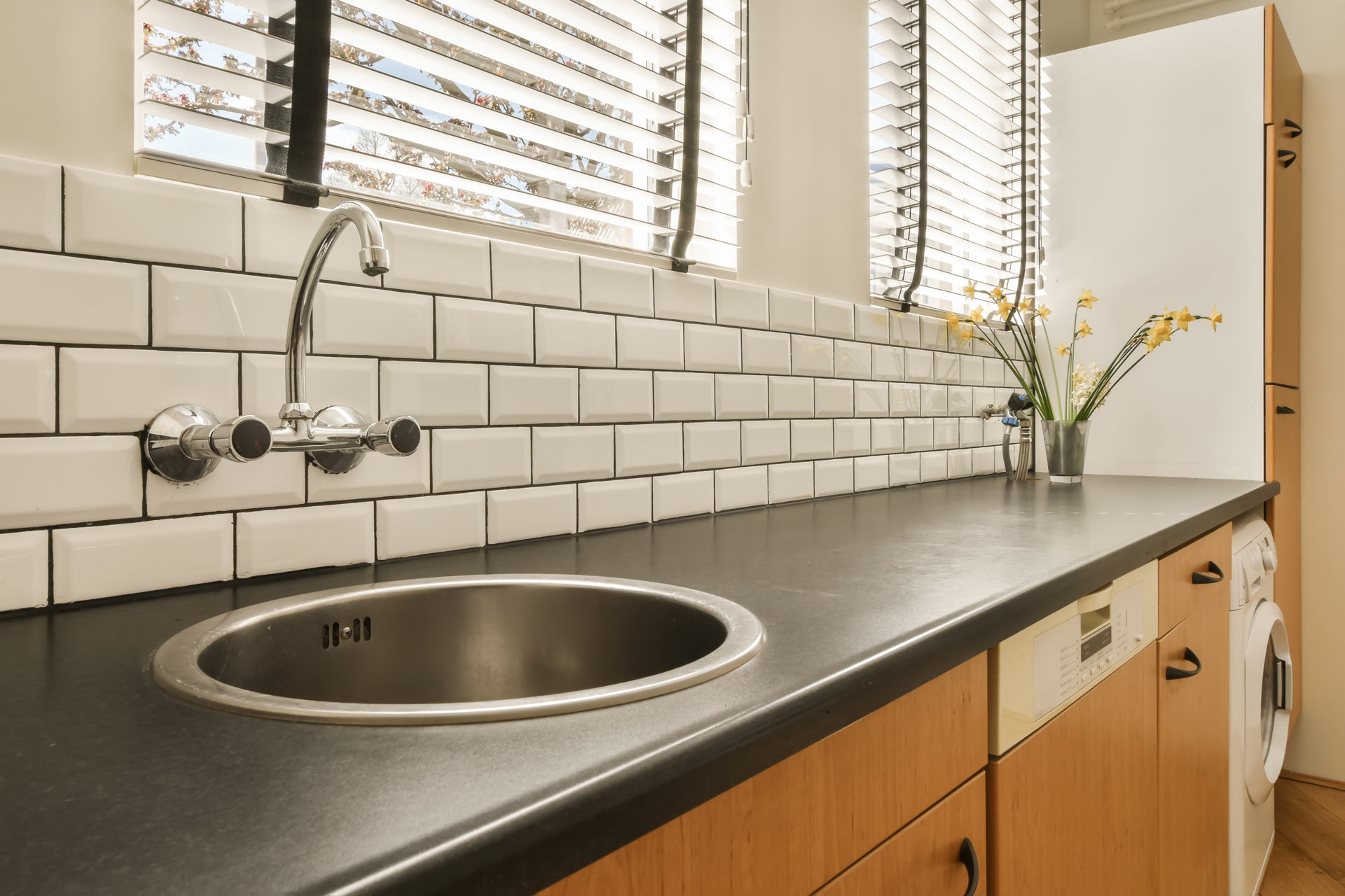 Plan de travail de cuisine avec évier rond en inox, robinet mural chromé, crédence en carreaux blancs et meubles en bois clair, éclairé par une fenêtre avec stores.