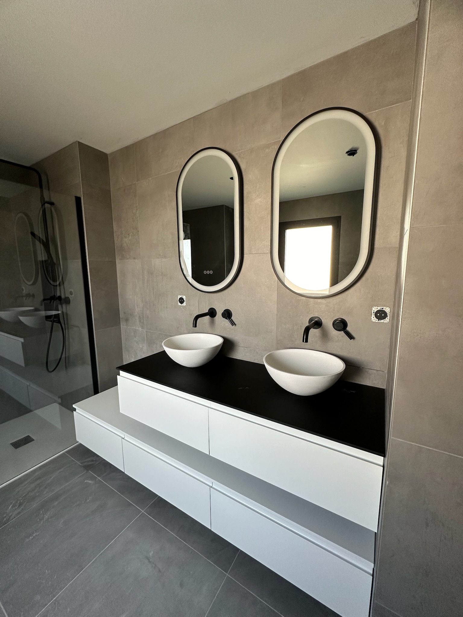 Plan de travail de cuisine avec évier rond en inox, robinet mural chromé, crédence en carreaux blancs et meubles en bois clair, éclairé par une fenêtre avec stores.